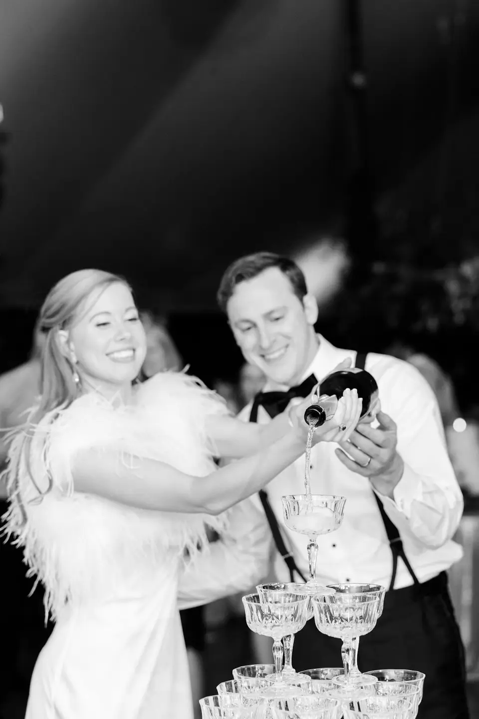 Black and white photo of a bride and groom pouring champagne over a tower of coupe glasses