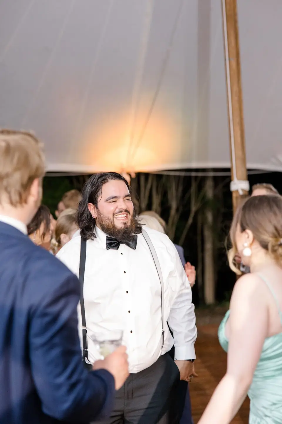 Smiling man in a bow tie and suspenders talking with other guests under a tent at a wedding