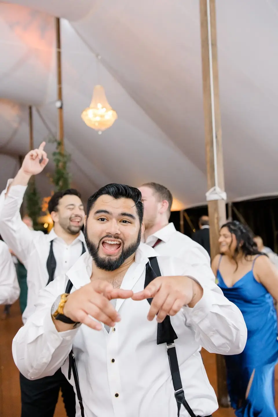 Man in a white shirt and suspenders pointing at the camera and cheering while guests dance behind him