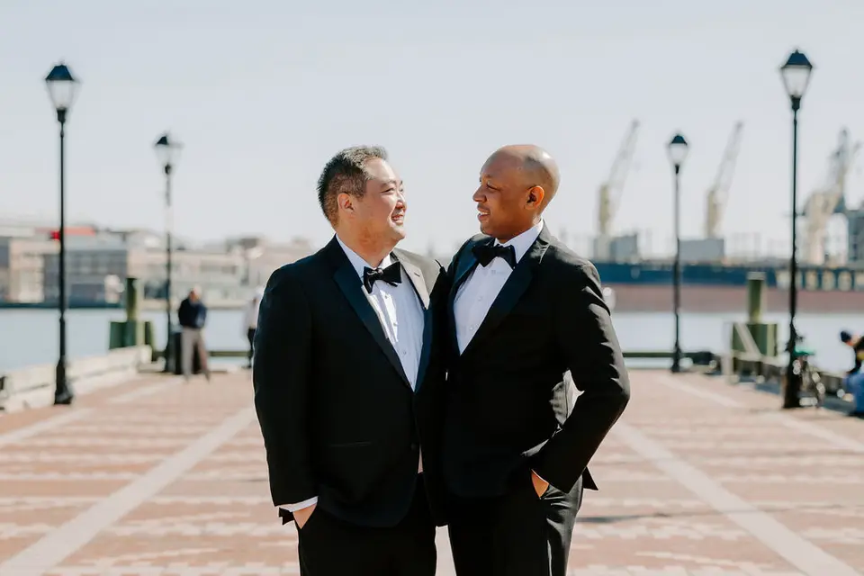 Two grooms in black tuxedos and bow ties smiling at each other on a waterfront promenade with cranes behind