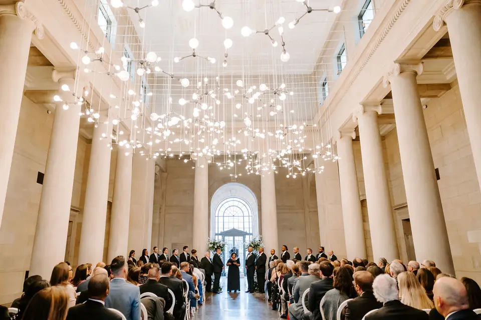 Wedding ceremony guests seated in a grand columned hall with hanging lights and a couple at the far end