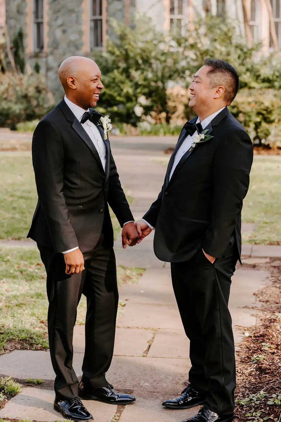 Two grooms in tuxedos holding hands and laughing on a stone path with boutonnieres on their lapels