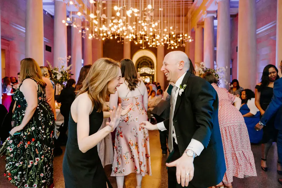 Guests dancing in a columned venue under a hanging light installation, a man in a tuxedo smiling in the center