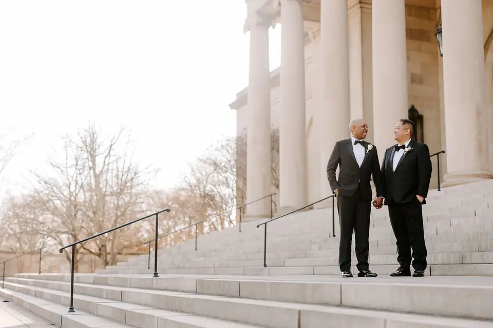 Two grooms in tuxedos standing on wide stone steps in front of a columned building