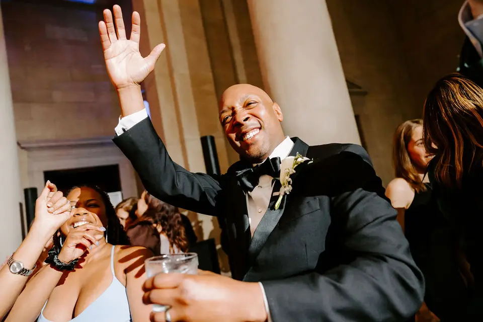 Smiling groom in a tuxedo and bow tie raising a hand and holding a drink on the dance floor
