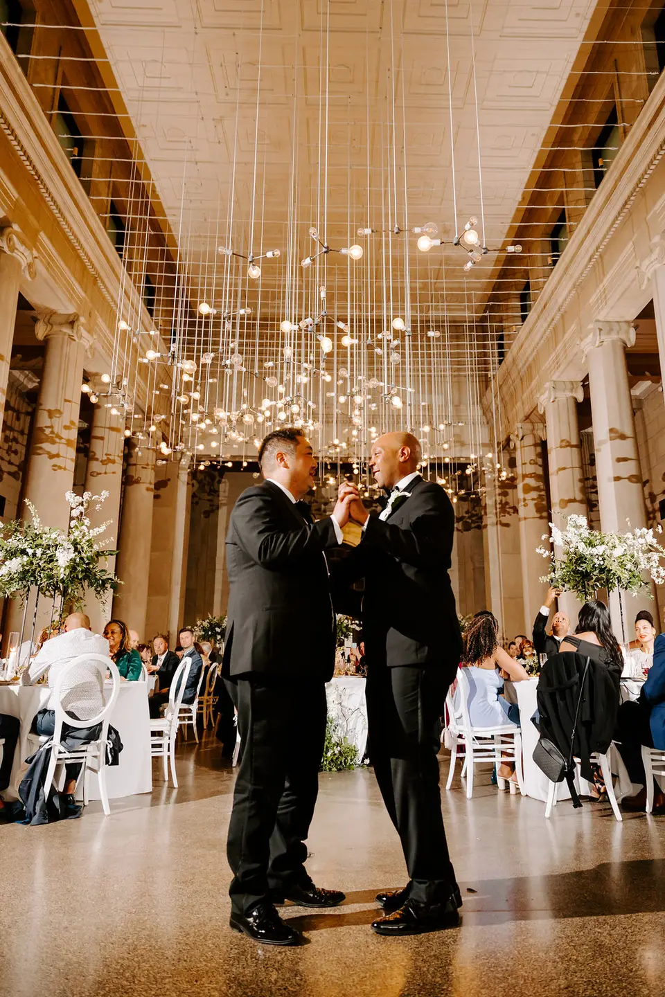 Two grooms dancing together in a large columned hall with strands of hanging lights overhead and guests seated nearby