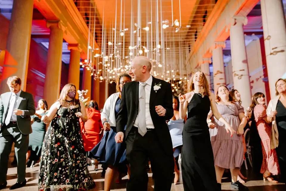 Groom in a tuxedo dancing with guests in a columned hall with hanging lights and warm colored uplighting