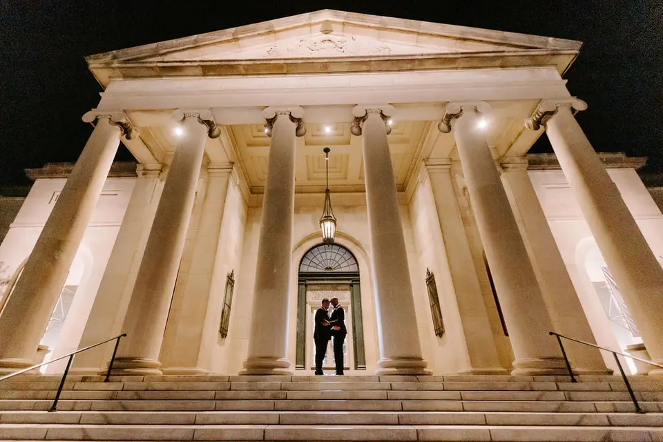 Two grooms standing at the entrance of a columned building at night, lit from below