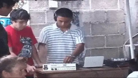 Young man in a striped shirt and headphones mixing on a laptop and controller outdoors as others watch