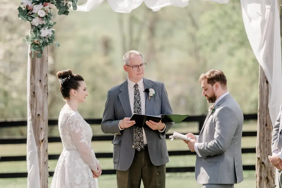 Officiant reads to the bride and groom during an outdoor ceremony under a draped wooden arbor