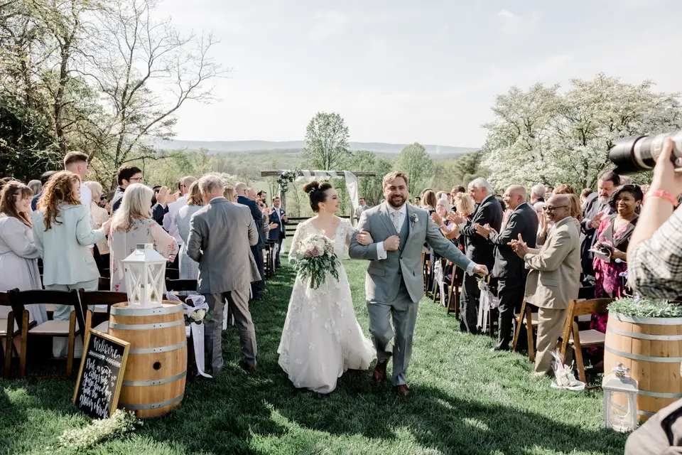 Newlyweds walk back up the grass aisle holding hands as guests applaud on a hillside