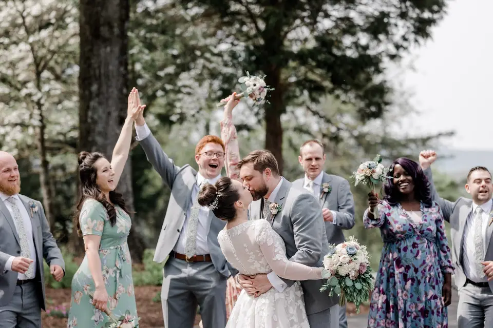 Bride and groom kiss while bridesmaids and groomsmen cheer with raised arms and bouquets outdoors