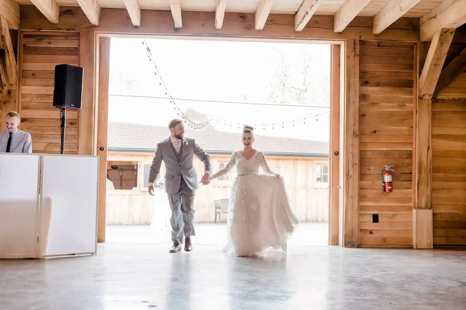 Bride and groom enter a rustic barn hand in hand while a DJ stands behind a white booth