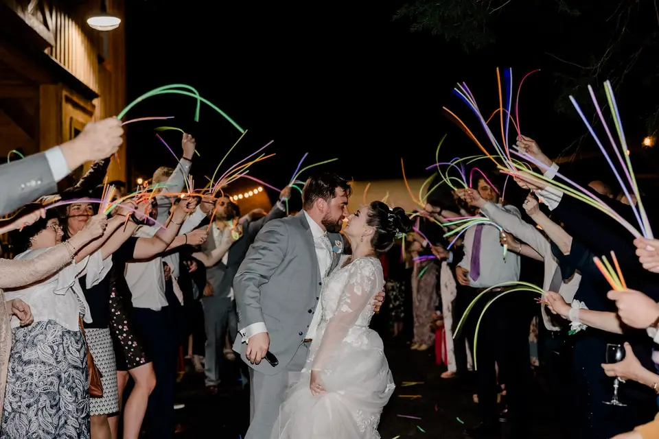 Bride and groom kiss at night between two lines of guests holding glow sticks