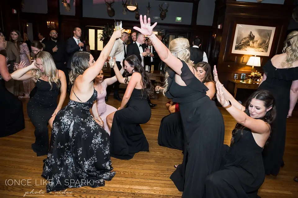 Bridesmaids in black dresses dance with raised arms on a wood dance floor in a paneled room