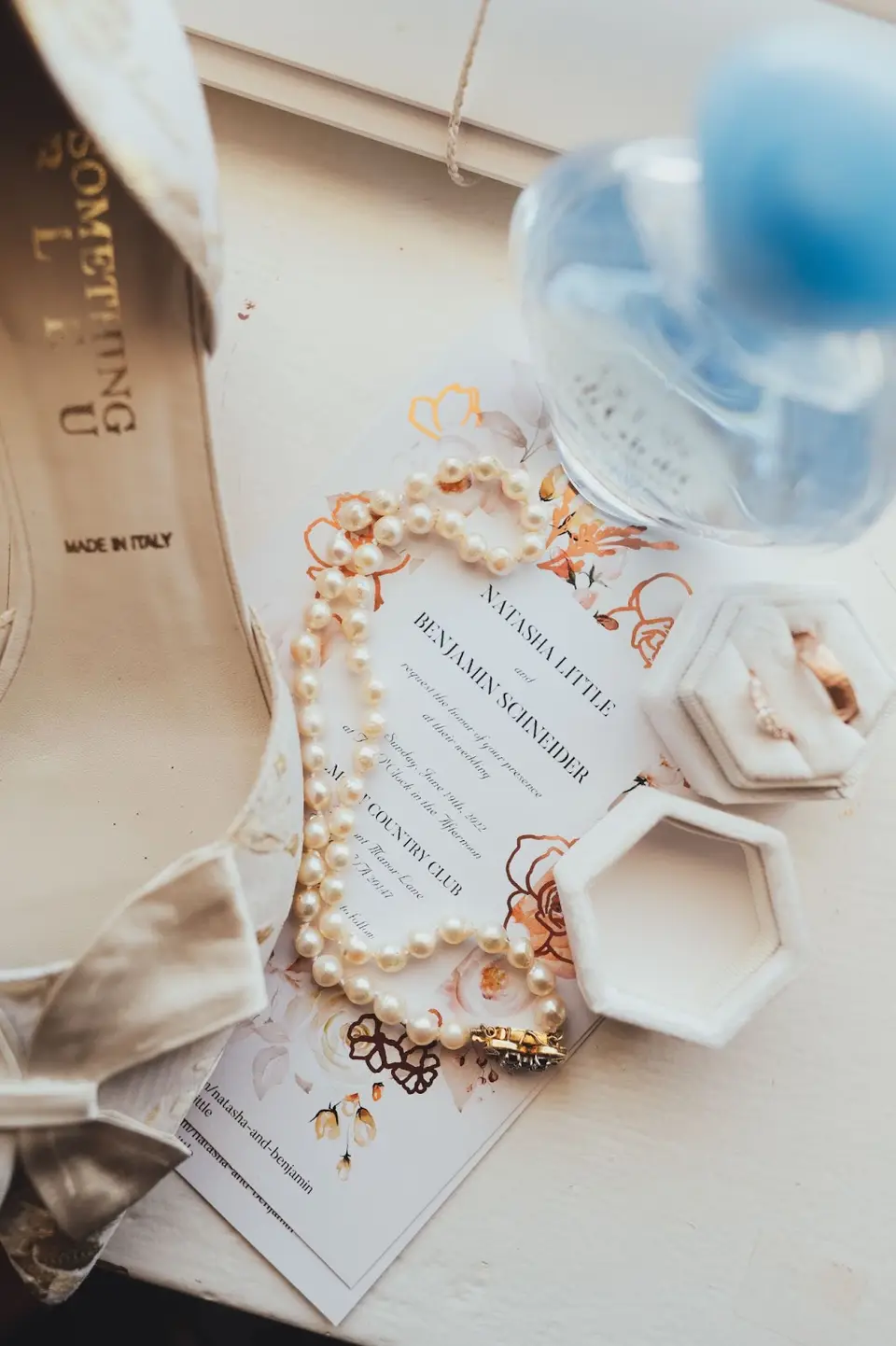 Bridal flat lay with a wedding invitation, pearl necklace, ring boxes, perfume bottle and blue-detailed bridal shoe