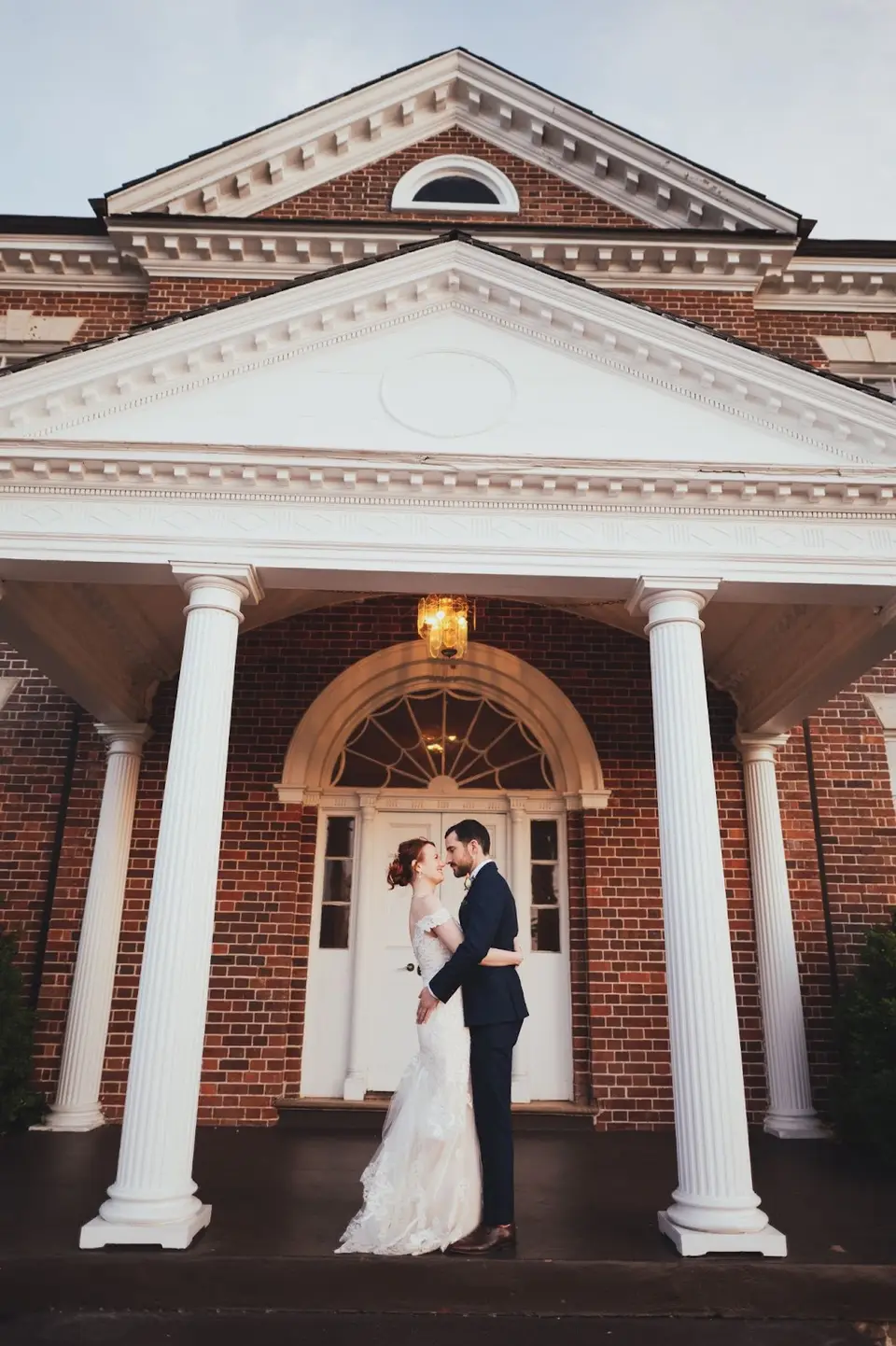 Bride and groom embracing under the white-columned portico of a brick country club entrance