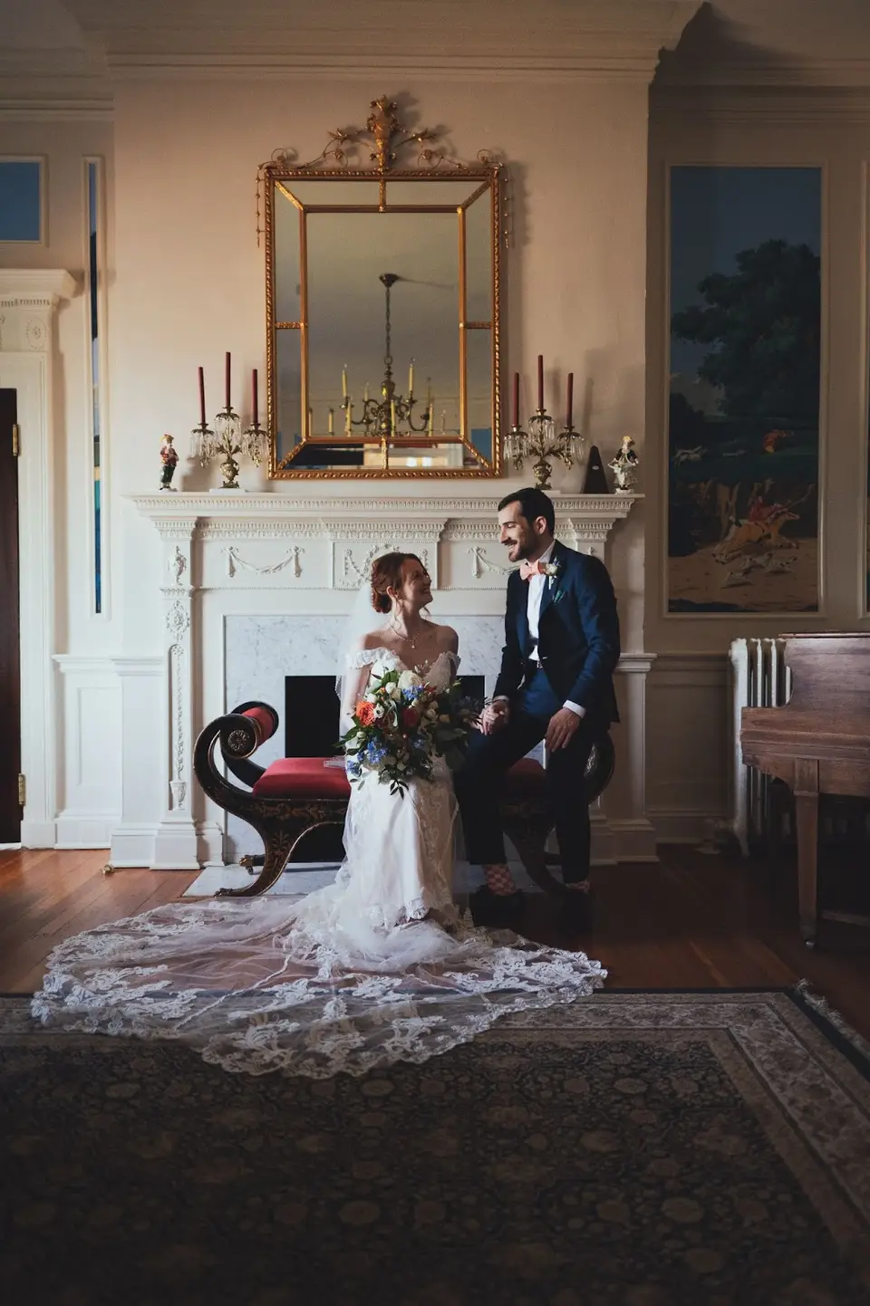 Bride and groom seated on a red bench in a formal room with a fireplace, mirror and candlesticks