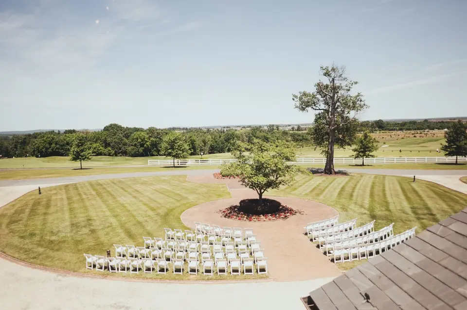 Rows of white chairs set for an outdoor ceremony on a manicured lawn around a tree