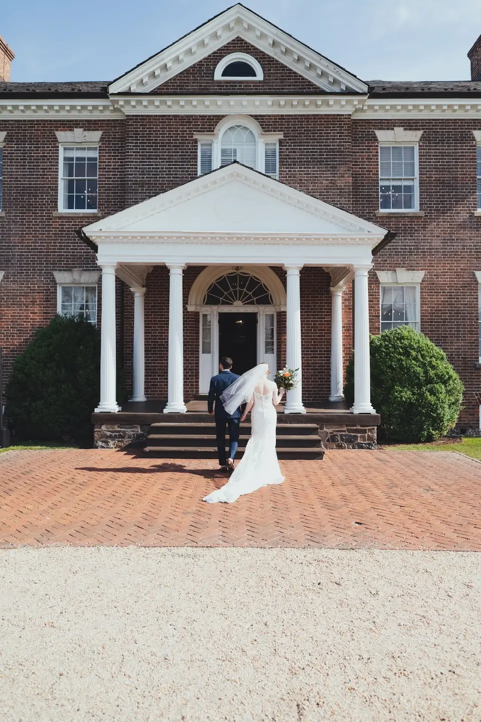 Bride in a veil and groom in a navy suit walking toward a red brick colonial building with white columns
