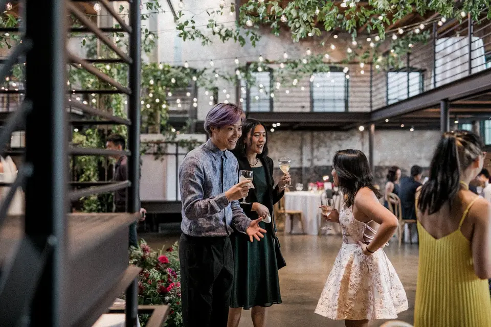 Guests chat with drinks at a cocktail hour in a brick industrial venue hung with string lights and greenery