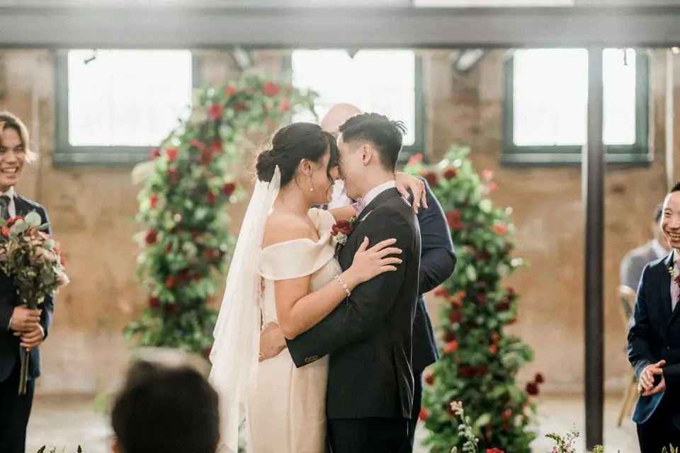 Bride and groom embrace at the ceremony in front of arches of green leaves and red roses