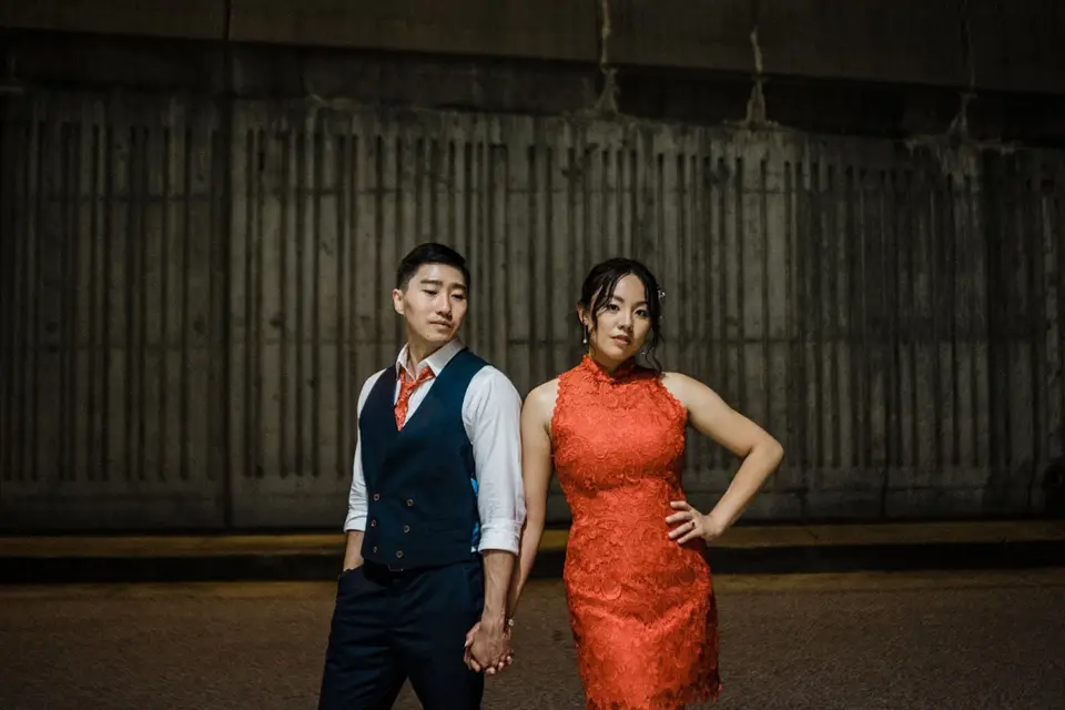 Couple in coordinated outfits, she in an orange lace dress and he in a navy vest, pose in front of a concrete wall