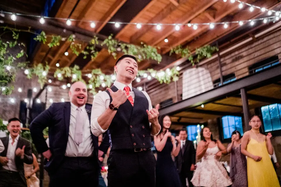 Smiling groom in a navy vest and red tie gestures to the crowd beneath string lights