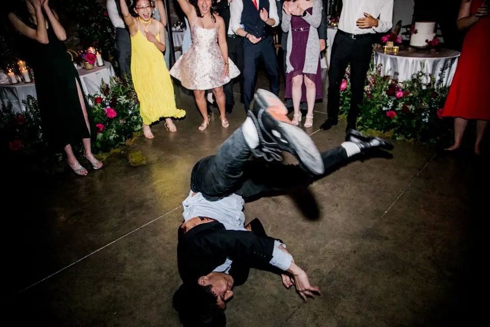 Man in a dark shirt flips on the dance floor while guests in dresses and suits watch