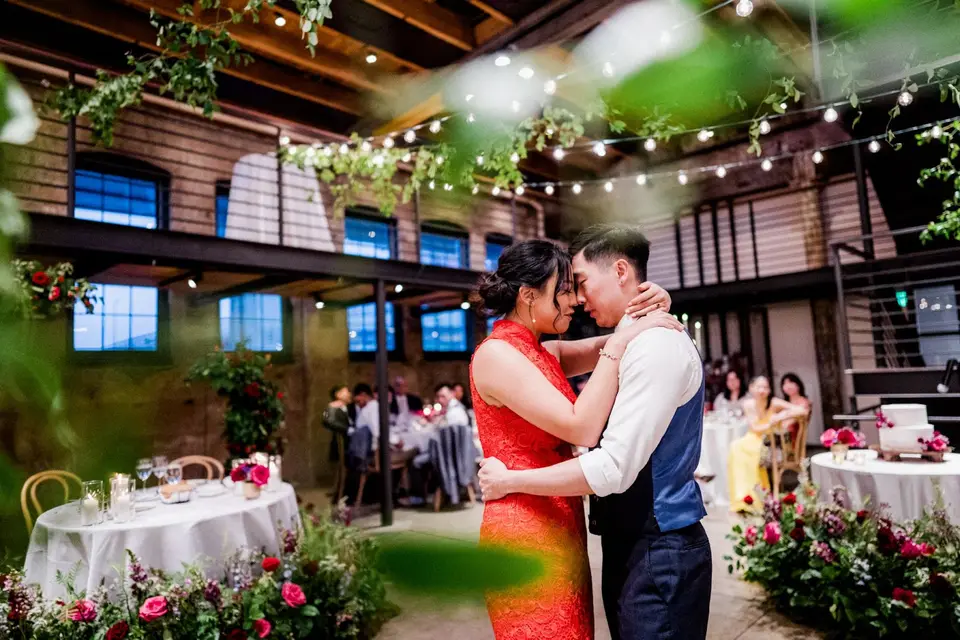 Bride in an orange dress and groom share a slow dance among flowers in a brick venue with string lights