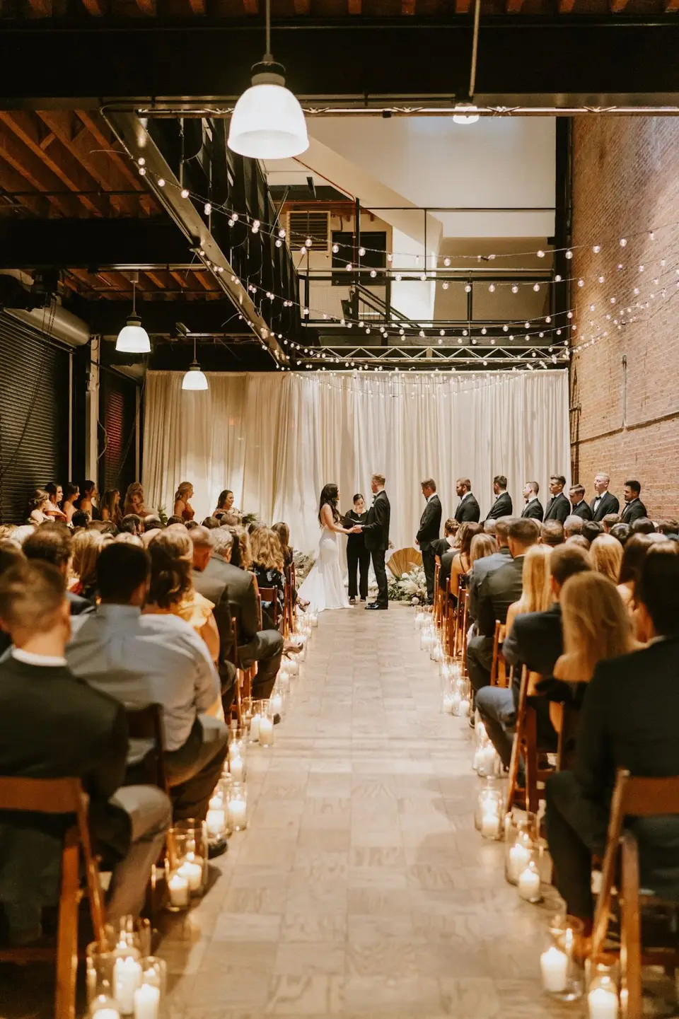 Wedding ceremony in an industrial brick venue with the couple at the end of a candlelit aisle lined with guests