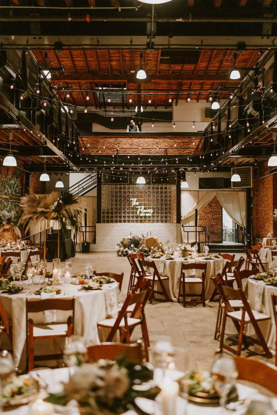 Reception tables with greenery centerpieces and wooden chairs in a brick industrial hall under string lights