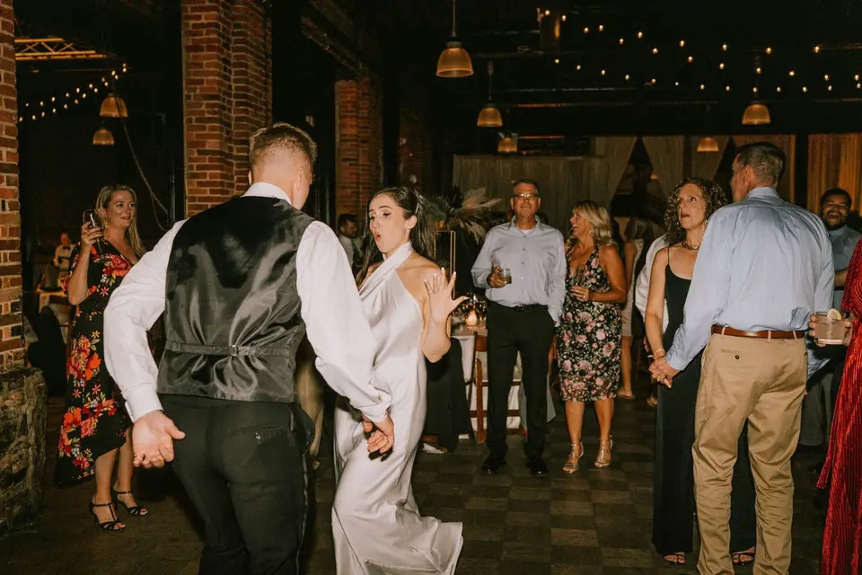 Bride in a satin gown and groom in a vest dancing together among reception guests