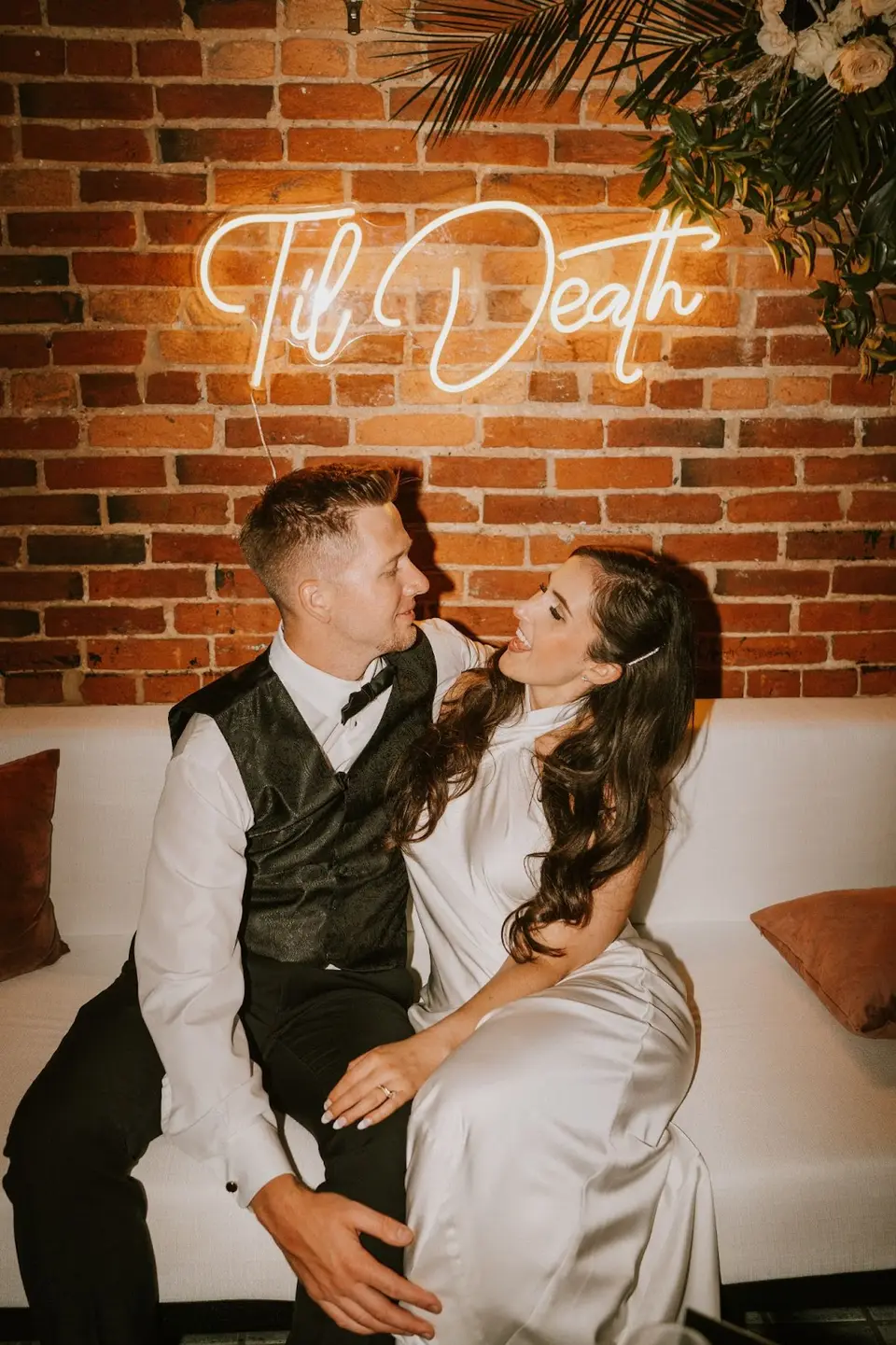 Bride and groom seated on a white couch beneath a neon sign reading Til Death
