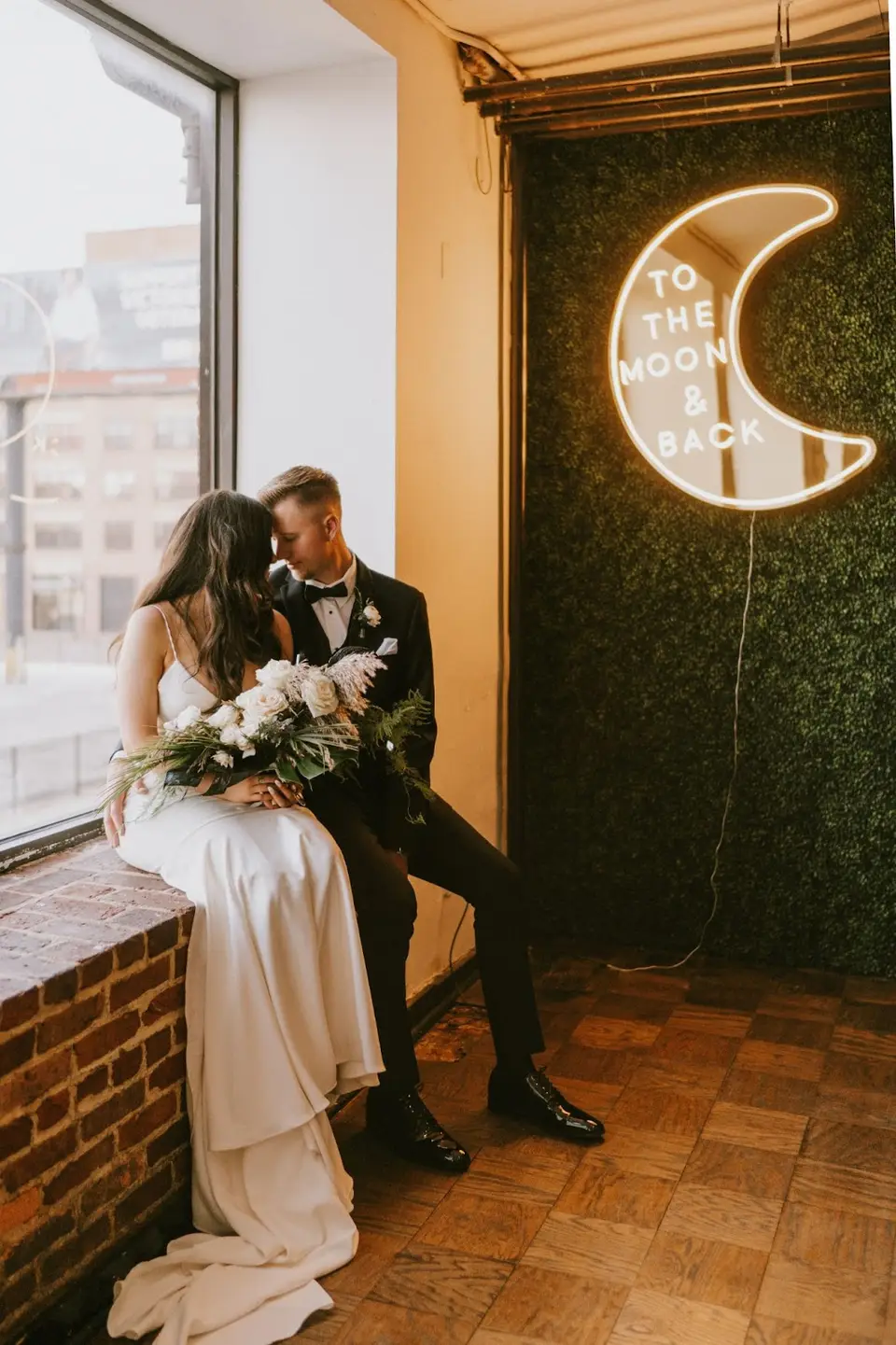 Bride and groom seated by a window beside a neon crescent moon sign reading To the Moon and Back