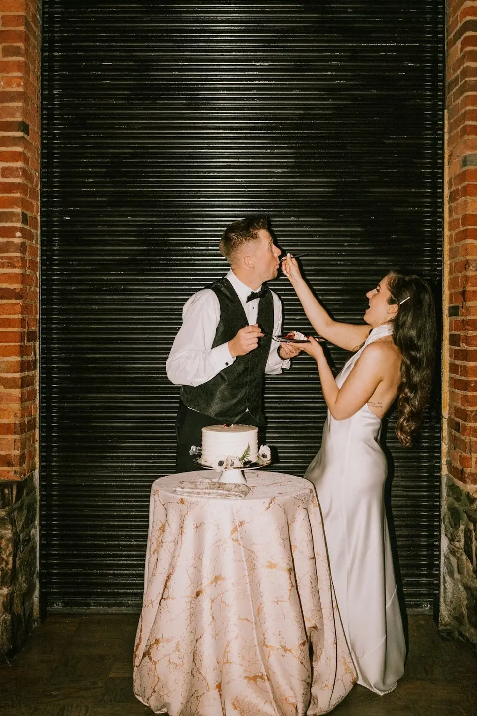 Bride feeding the groom a bite of cake at a small table in front of a black roll-up door