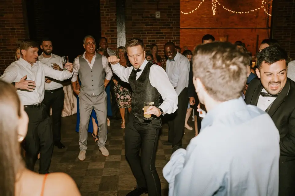 Groom in a vest raising a drink and dancing with guests during the reception