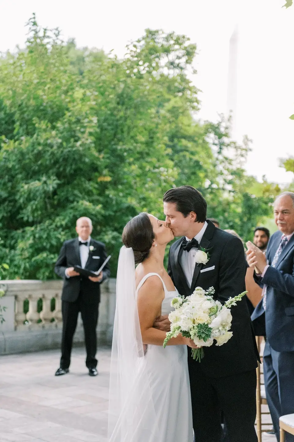 Bride and groom kissing at the end of an outdoor ceremony while the officiant stands behind them