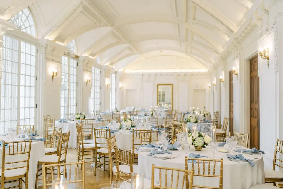 Ballroom with arched ceiling and tall windows set with round tables, gold chairs and white and blue florals
