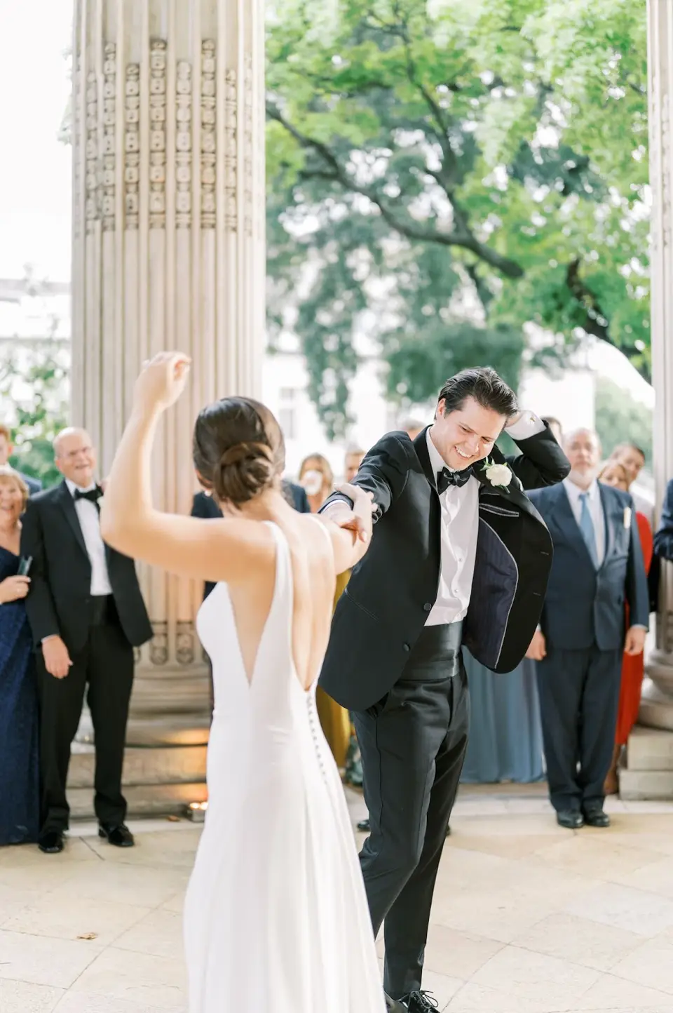 Groom taking off his tuxedo jacket while the bride dances with her arm raised between stone columns