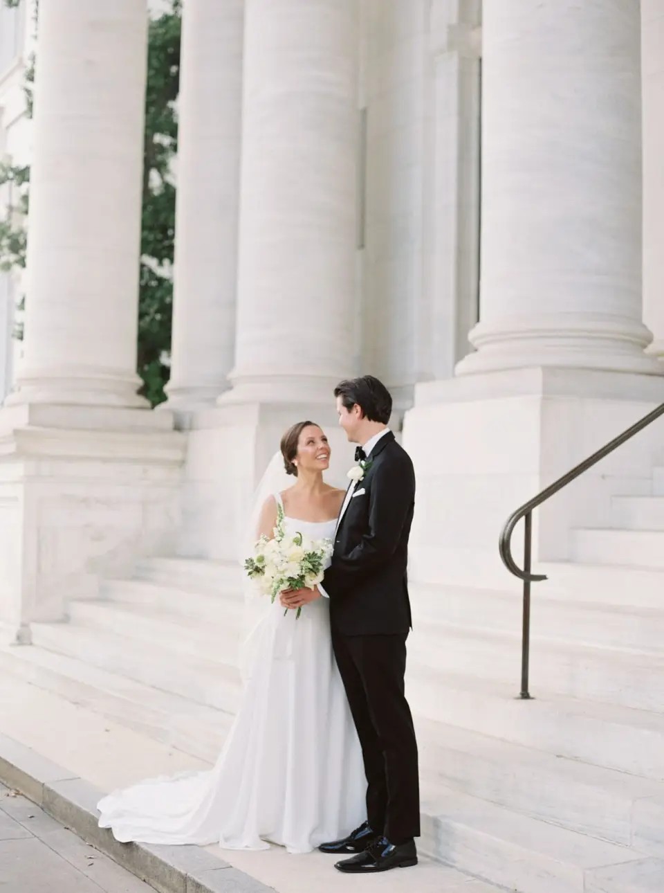 Bride holding a white bouquet and groom in a black tuxedo posing on stone steps beside large columns