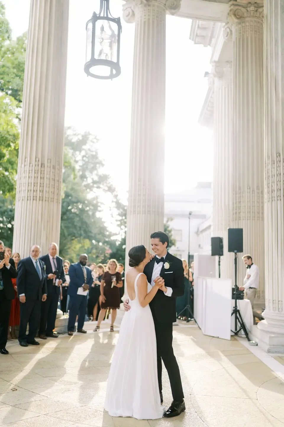 Bride and groom dancing between tall stone columns as guests look on and speakers stand nearby