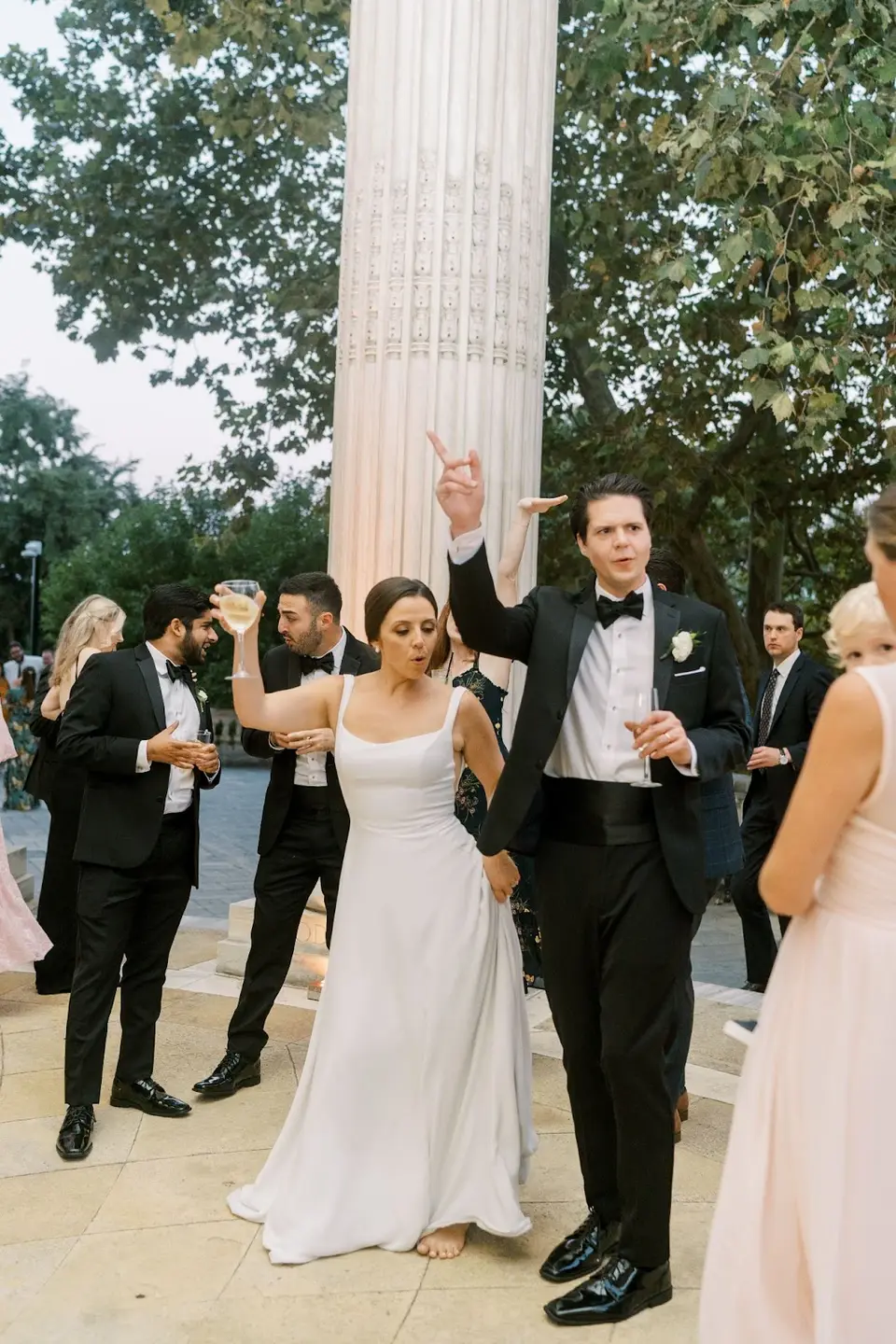 Groom with a raised arm dancing with the bride among guests on a stone terrace outdoors