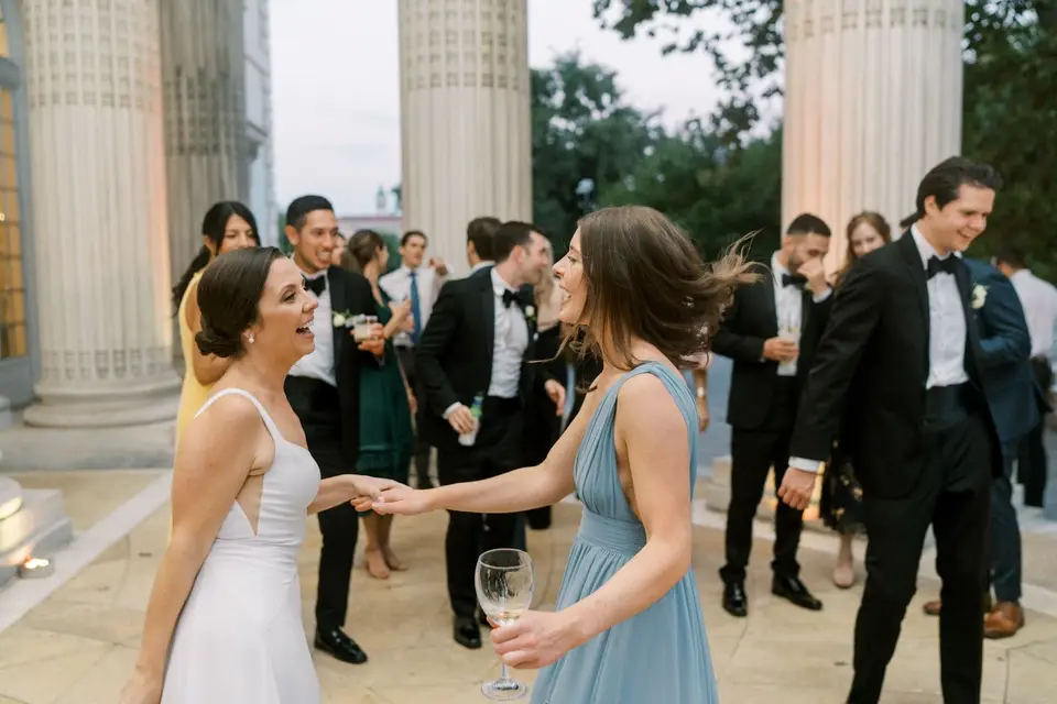 Guests laughing and dancing on a stone terrace between columns, including a bridesmaid in light blue