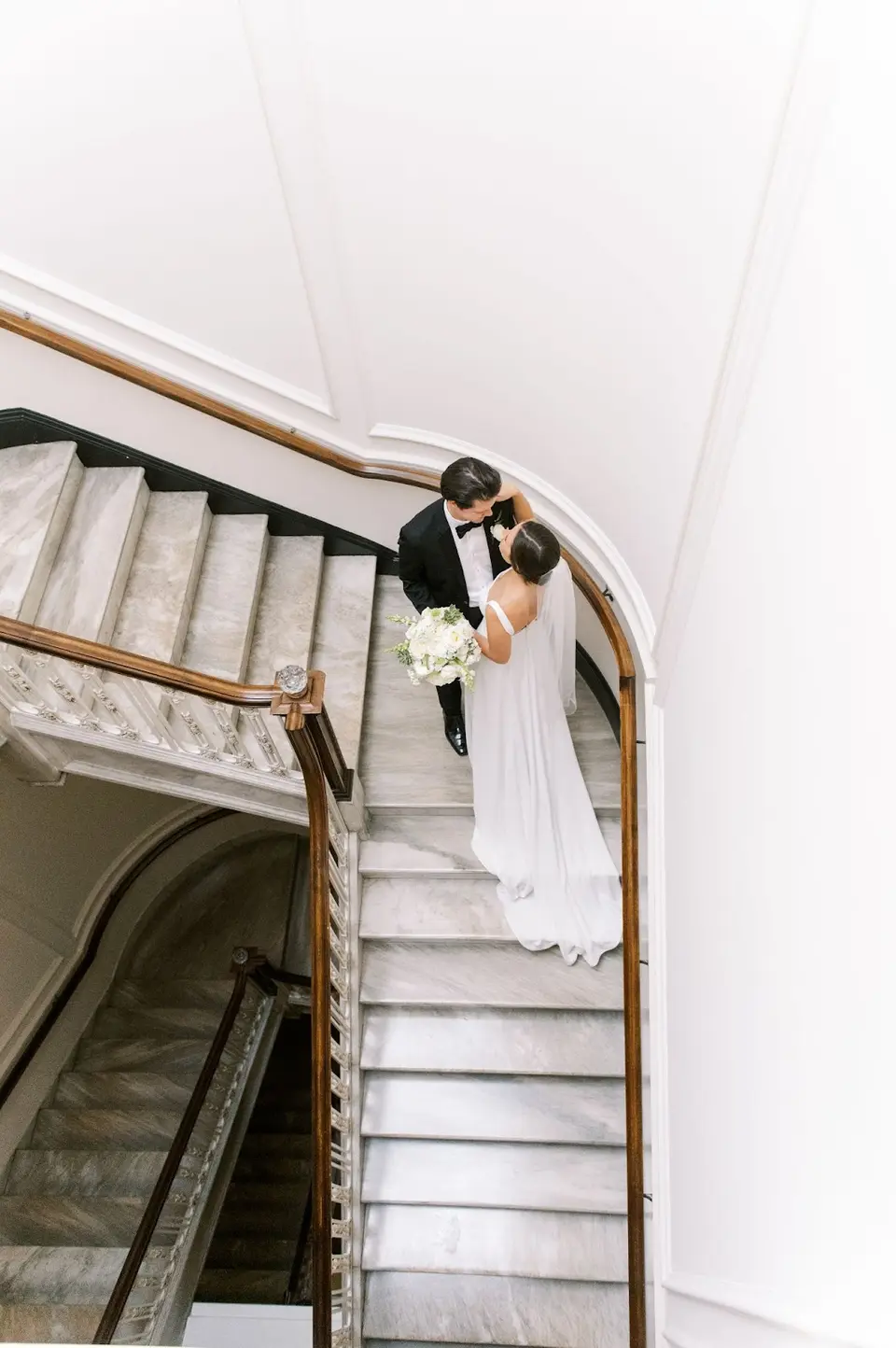 Bride and groom embracing on a marble staircase, seen from above, holding a white bouquet