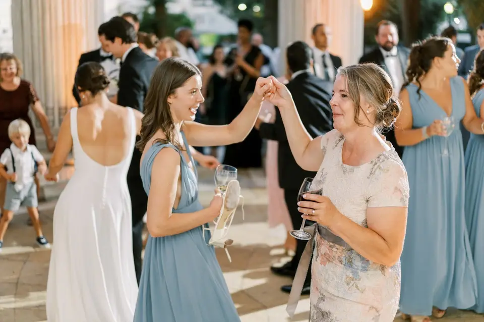 Two women in blue dresses dancing and holding hands with other guests on an outdoor terrace