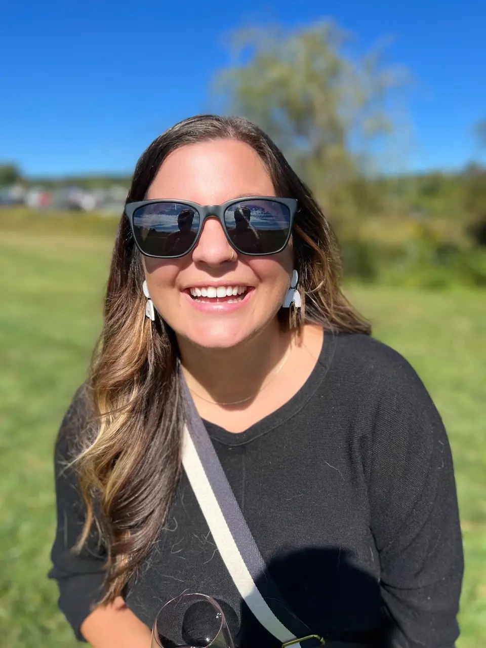 Smiling woman with long hair and sunglasses outdoors on a grassy field under a blue sky