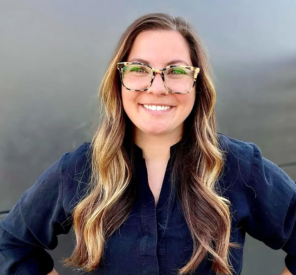 Smiling woman with long highlighted hair and tortoiseshell glasses, hands on hips, against a gray wall
