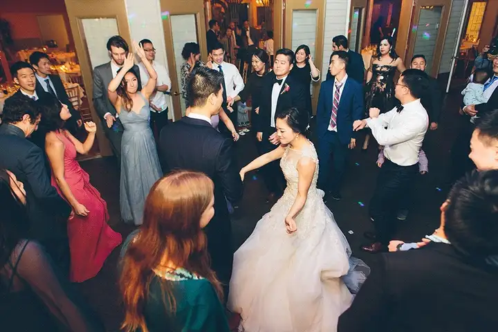 Bride in a white gown and guests dancing and clapping together on a crowded wedding dance floor