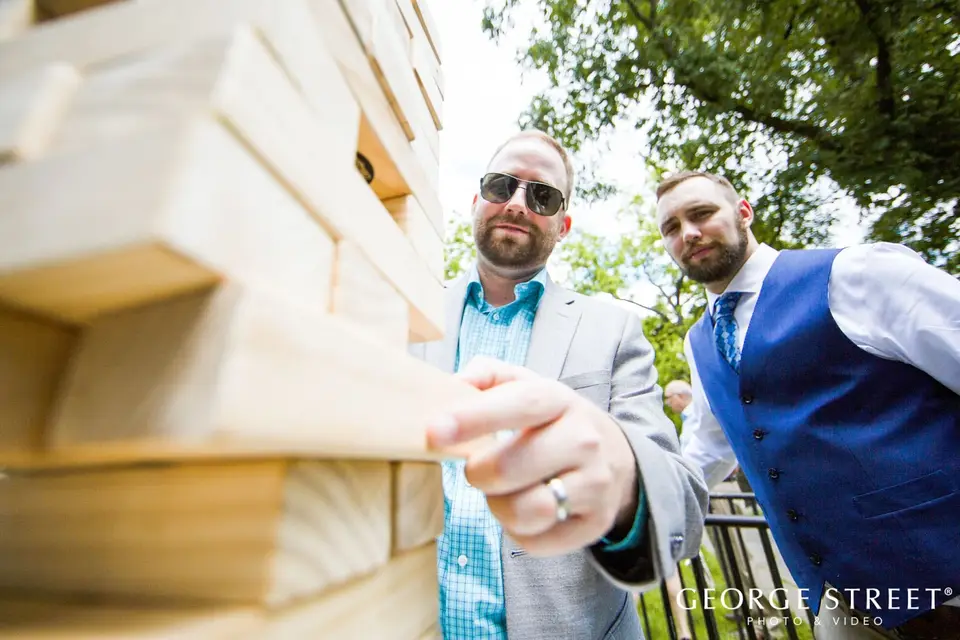 Two men in vests and sunglasses leaning over a giant wooden Jenga tower at an outdoor wedding